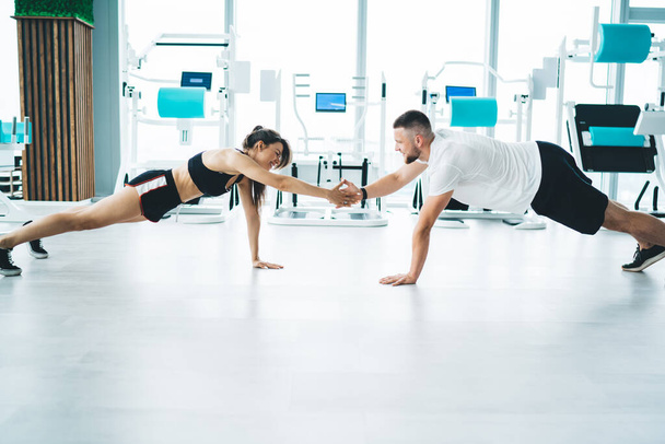 Side view of young content slim sportspeople touching hands while standing in plank on floor and looking at each other in gymnasium - Фото, изображение