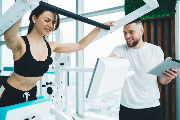 Crop young male trainer with tablet talking to female athlete in sportswear exercising on modern electronic equipment in light gymnasium - Foto, Imagen