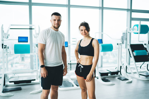 Young sportsman near content female partner in active wear standing with hand on waist against exercise machines while looking at camera in gymnasium - Foto, Bild