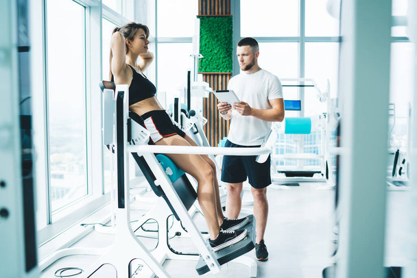 Bearded trainer with tablet near female athlete in sportswear working out with hands behind head on electronic machine in gymnasium - Fotoğraf, Görsel