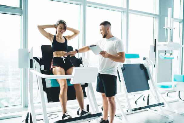 Young content male coach with tablet touching abs of sportswoman in active wear exercising with hands behind head on modern fitness machine - 写真・画像