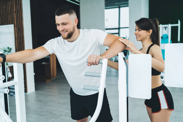 Positive fit male athlete improving endurance and vitality while exercising on sports simulators in modern gym with young female instructor - Foto, Imagen