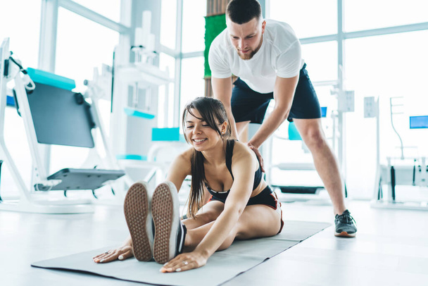 Diligent cheerful young sportswoman in activewear stretching on floor with experienced trainer in gym with large windows and gym equipment - Foto, Bild