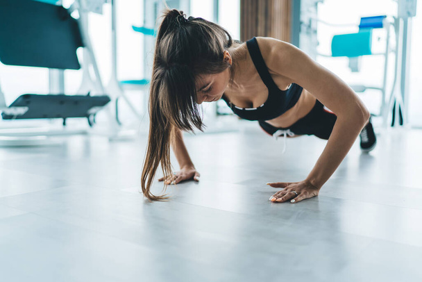 Focused young slim female in activewear doing push ups on floor on blurred background of modern comfortable gym with equipment - Φωτογραφία, εικόνα