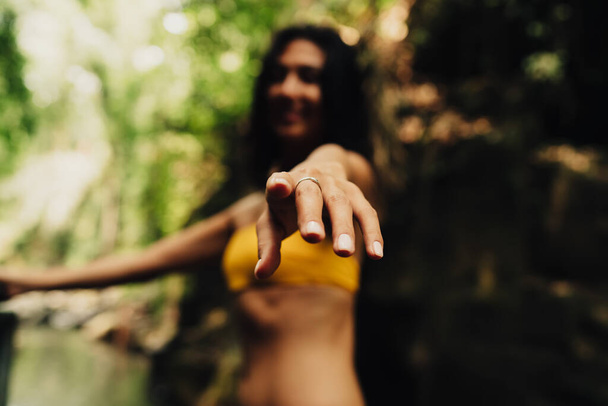 Happy anonymous female in bright yellow bikini holding hand out in green forest on warm sunny day in soft focus - Zdjęcie, obraz