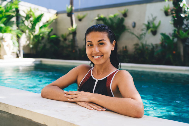 Young attractive ethnic woman with toothy smile leaning on edge of swimming pool surrounded by greenery and concrete walls of hotel yard - Fotó, kép