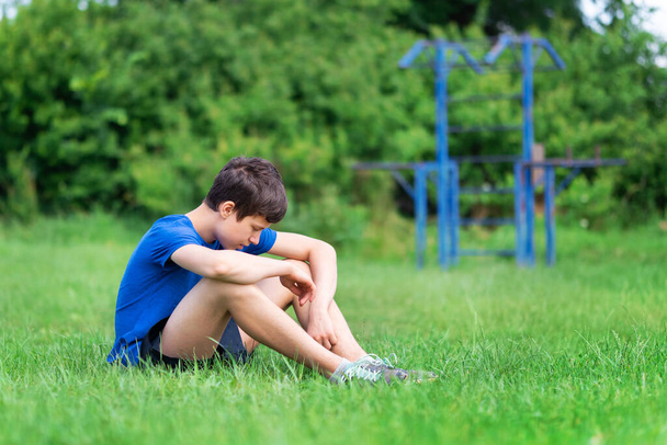 teenage boy exercising outdoors, sports ground in the yard, he sits on the green grass of the playground, healthy lifestyle - Foto, Imagen