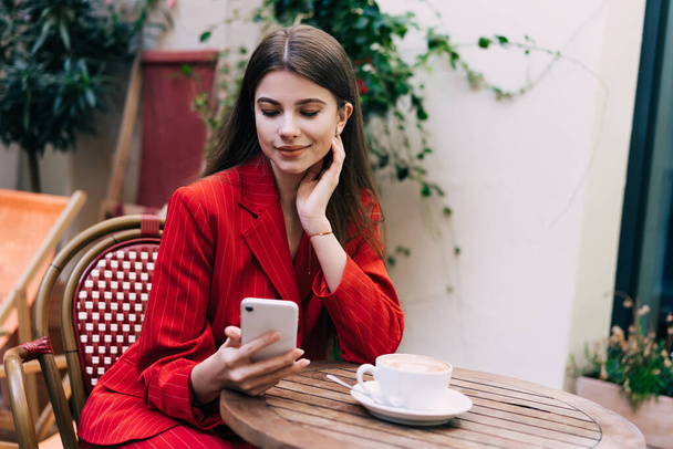 Happy young female in stylish outfit messaging on cellphone while smiling and sitting at table with hand on chin pose at cozy cafe - Foto, Imagem