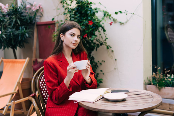 Young female in trendy outfit smiling and sitting at round wooden table while holding cup of coffee in hands outside cafe - Фото, зображення
