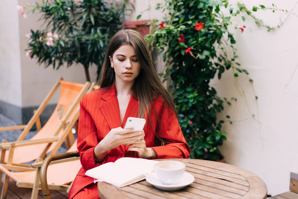 Serious businesswoman in stylish clothes sitting at small round table in street cafe and browsing mobile phone while working on project - Photo, image
