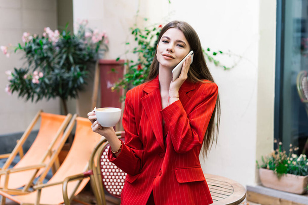 Ευτυχισμένη νεαρή γυναίκα με κομψά ρούχα μιλάει στο κινητό τηλέφωνο, ενώ στέκεται στη βεράντα στο δρόμο καφέ και πίνοντας φλιτζάνι καφέ - Φωτογραφία, εικόνα