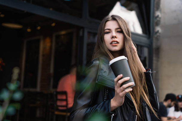 Stunning stylish lady in black leather jacket holding cup of takeaway hot drink and looking at camera while standing near entrance of coffee shop - Fotoğraf, Görsel