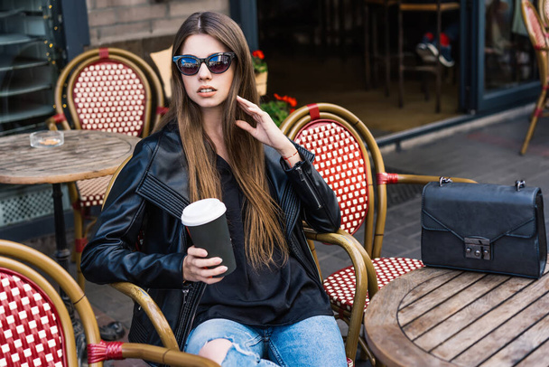 Stylish female model in fashionable outfit and sunglasses holding cup of coffee in hand and looking away while sitting at table with handbag and posing with legs crossed - Photo, Image