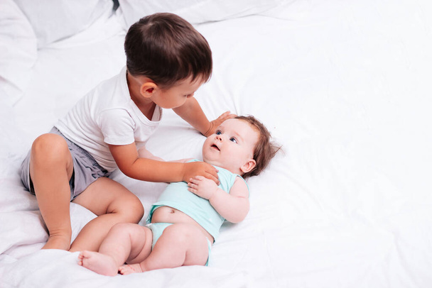 симпатичный мальчик обнимает младшую сестру на белоснежной кровати - Фото, изображение