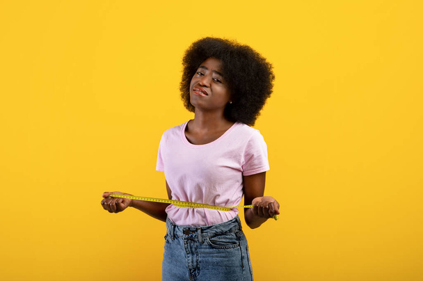 Gewichtsverlust und Schlankheitskonzept. Enttäuschte afrikanische Amerikanerin misst Taille mit Klebeband, gelber Hintergrund - Foto, Bild