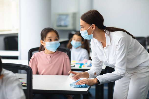 Teacher in mask disinfecting table with antibacterial sanitizer and napkin - Photo, Image