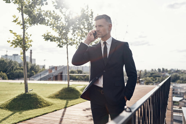 Confident mature businessman talking on mobile phone while walking outdoors - Foto, Bild