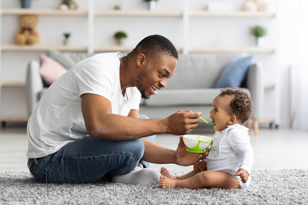 Fathers Care. Loving Black Dad Feeding His Cute Baby Son At Home - Zdjęcie, obraz