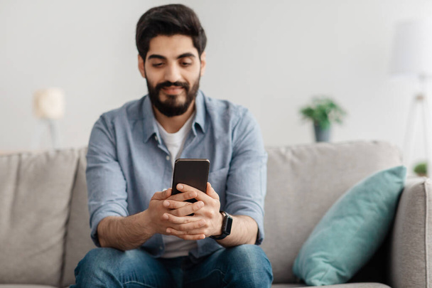 zbliżenie arabski facet w dorywczy strój za pomocą nowoczesny smartfon, odpoczynek w domu wnętrze, kopia przestrzeń - Zdjęcie, obraz