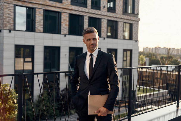 Confident mature man in full suit carrying laptop while standing on business center terrace - Fotoğraf, Görsel