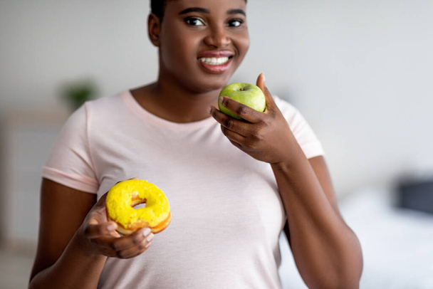 Donne afro paffute che scelgono tra mela e ciambella, selezionando frutta, preferendo un'alimentazione più sana per la perdita di peso - Foto, immagini