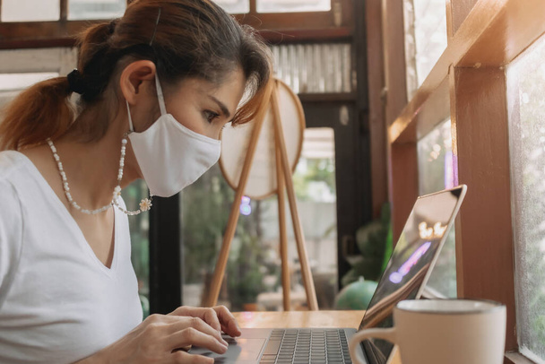 Γυναίκα με μάσκα προσώπου εργάζεται σε φορητό υπολογιστή στο καφέ. - Φωτογραφία, εικόνα