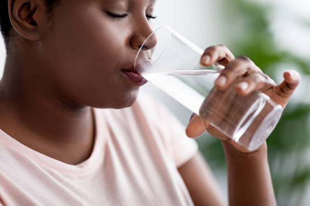 Поддерживайте гидратацию для более здорового образа жизни. Избыточный вес черная женщина пьет чистую минеральную воду из стекла в помещении - Фото, изображение