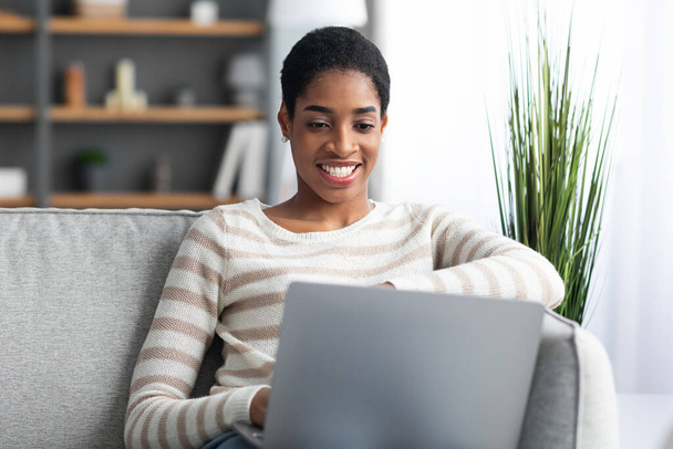 Uzaktan kumanda. Gülümseyen Siyah Bağımsız Kadın Koltukta otururken Dizüstü bilgisayar kullanıyor - Fotoğraf, Görsel
