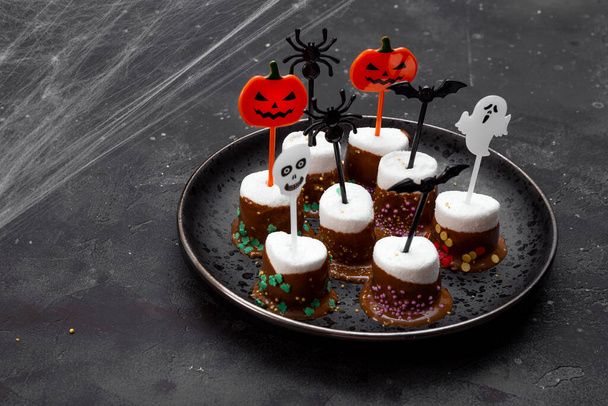  Sweet Halloween treat, chocolate covered marshmallows with sugar and Halloween decorations on a black plate, photo with soft focus and free space for text - Fotó, kép