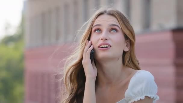 Emotional female student in summer dress share amazing news with friend by phone hold device by ear. Surprised young lady answer telephone call to hear about winning lottery getting unexpected reward - Footage, Video