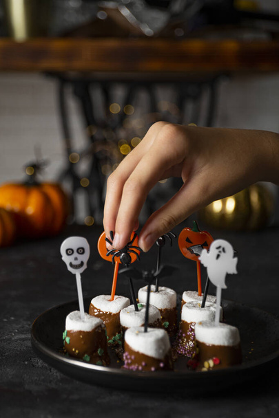  Sweet Halloween treat, chocolate covered marshmallows with sugar and Halloween decorations on a black plate, vertical photo with soft focus - Fotó, kép