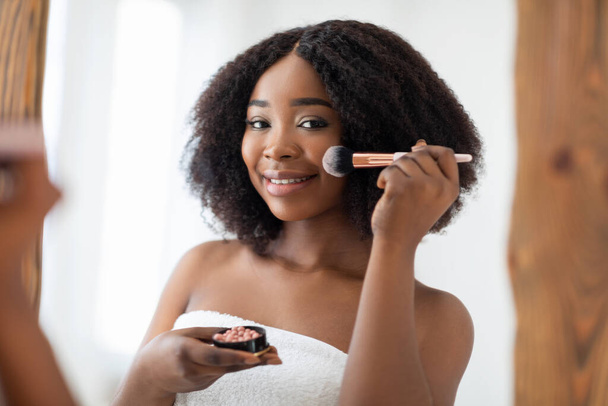 Koncept přirozeného make-upu. Okouzlující černošky dáma nanášení červenat nebo prášek před zrcadlem doma - Fotografie, Obrázek