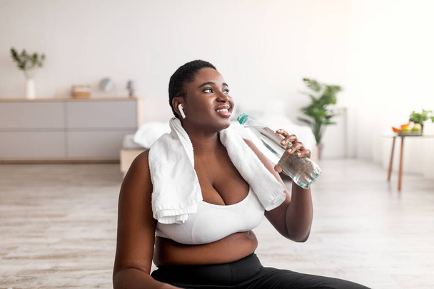Porträt einer jungen schwarzen Plus-Size-Frau mit Kopfhörern, die Wasser trinkt und in der Pause vom Heimtraining Musik hört - Foto, Bild