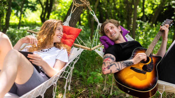 Couple lying on hammocks. Young man is playing guitar while woman is looking at him. Greenery around. Glamping - Photo, image