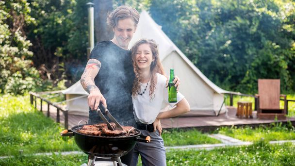 Young couple frying meat on the grill and drinking beer, laughing. Greenery around. Glamping - Photo, Image