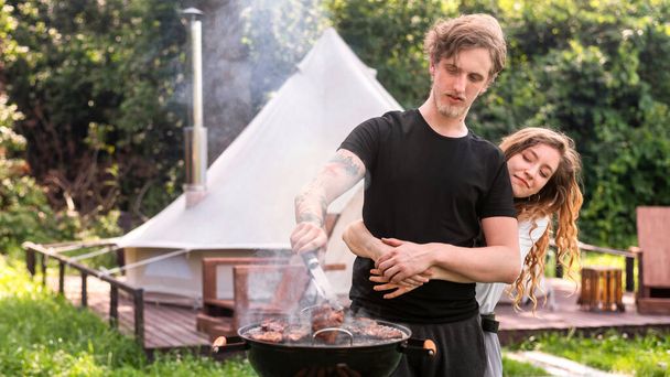 Young hugging couple frying meat on the grill. Greenery around. Glamping - Photo, Image