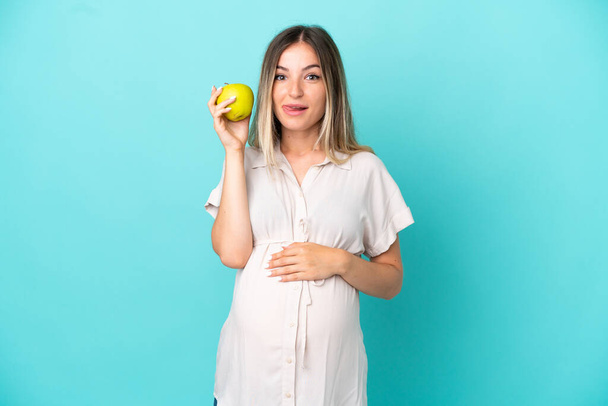 Joven rumana aislada sobre fondo azul embarazada y sosteniendo una manzana - Foto, Imagen
