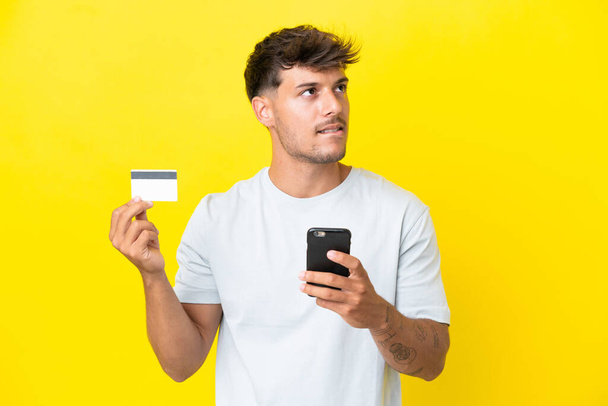 若いですcaucasianハンサムな男隔離上の黄色の背景購入とともにザ · 携帯電話とともにクレジットカード思考 - 写真・画像