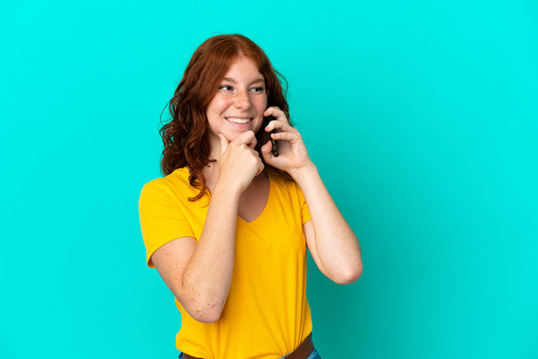 Mavi arka planda izole edilmiş cep telefonu kullanan genç kızılımsı bir kadın yana bakıyor ve gülümsüyor. - Fotoğraf, Görsel