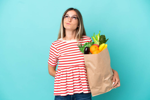 Junge Frau hält auf blauem Hintergrund eine Einkaufstasche in der Hand und schaut nach oben - Foto, Bild