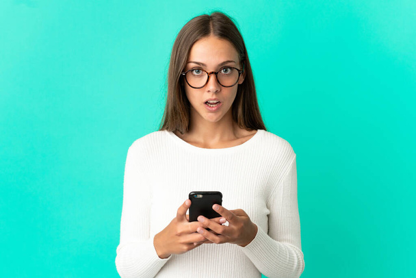 İzole edilmiş mavi arka planda genç bir kadın kameraya bakarken şaşırmış bir ifadeyle cep telefonuna bakıyor. - Fotoğraf, Görsel
