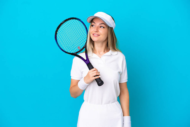 Młoda tenisistka Rumuńska kobieta odizolowana na niebieskim tle patrząc z boku i uśmiechnięta - Zdjęcie, obraz