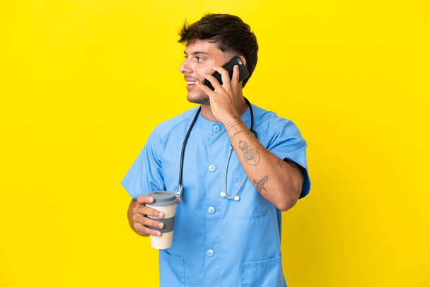 Nuori kirurgi lääkäri mies eristetty keltainen tausta tilalla kahvia ottaa pois ja mobiili - Valokuva, kuva