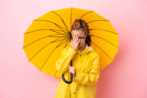 Teenager Russian girl with rainproof coat and umbrella isolated on pink background with tired and sick expression - Photo, Image