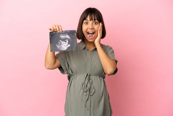 Frau über isolierten Hintergrund schwanger und überrascht, während sie einen Ultraschall hält - Foto, Bild