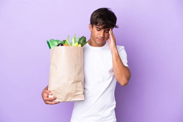 Νεαρός άνδρας κρατώντας μια τσάντα ψώνια απομονωμένη σε μωβ φόντο με πονοκέφαλο - Φωτογραφία, εικόνα