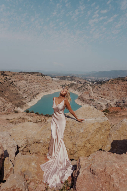 fashion outdoor photo of beautiful woman with blond hair in elegant dress posing on the top of mountain with lake view - Foto, imagen