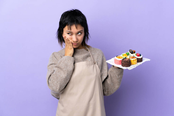 Giovane donna uruguaiana in possesso di un sacco di diverse mini torte su sfondo viola isolato infelice e frustrato - Foto, immagini