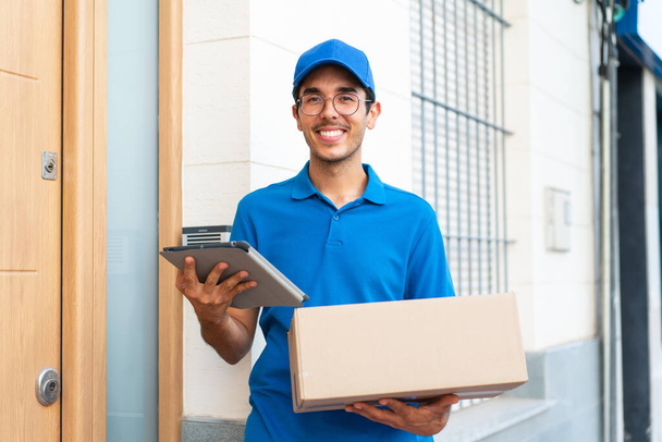 Молодой доставщик на открытом воздухе держит коробки и планшет с радостным выражением лица - Фото, изображение
