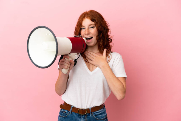 Teenager reddish woman isolated on pink background shouting through a megaphone with surprised expression - Valokuva, kuva